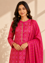 A woman modeling a bright pink traditional outfit with gold and white printed patterns, featuring a matching dupatta draped over her shoulder, set against a warm beige background with soft leaf shadows.