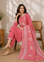 A woman seated on a wooden chair wearing a pink traditional outfit with matching pants and a long printed dupatta, photographed in a warm indoor setting with a side table and vase in the background.