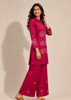 A woman modeling a magenta two-piece kurta set with white circular embroidery, standing against a solid beige studio background.