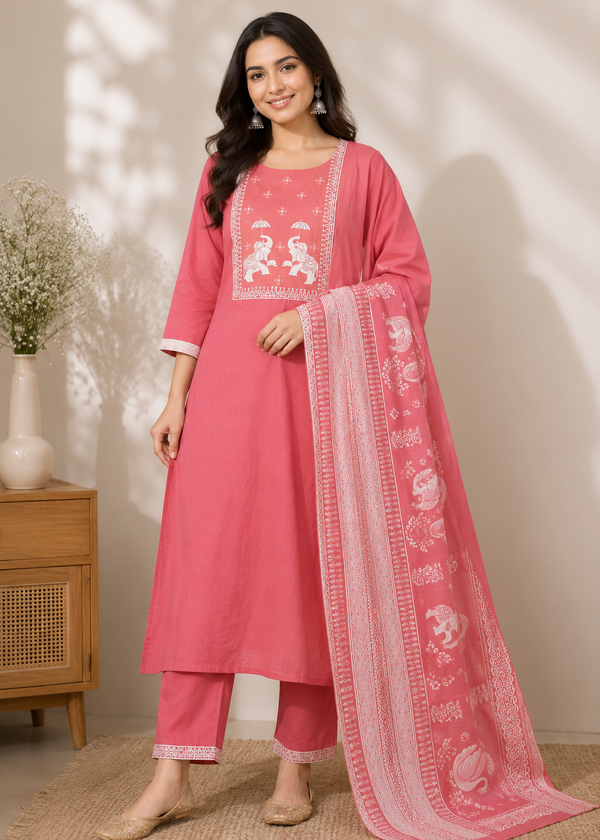 A woman models a pink Indian ethnic suit set consisting of a kurta, trousers, and a matching dupatta. The kurta features white embroidery on the chest, and the dupatta has white floral prints and borders.