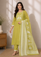 A woman models a three-piece lime green salwar kameez suit with white embroidery, featuring a long kurta, matching trousers, and a sheer printed dupatta, set against a warm beige wall with soft window shadows.