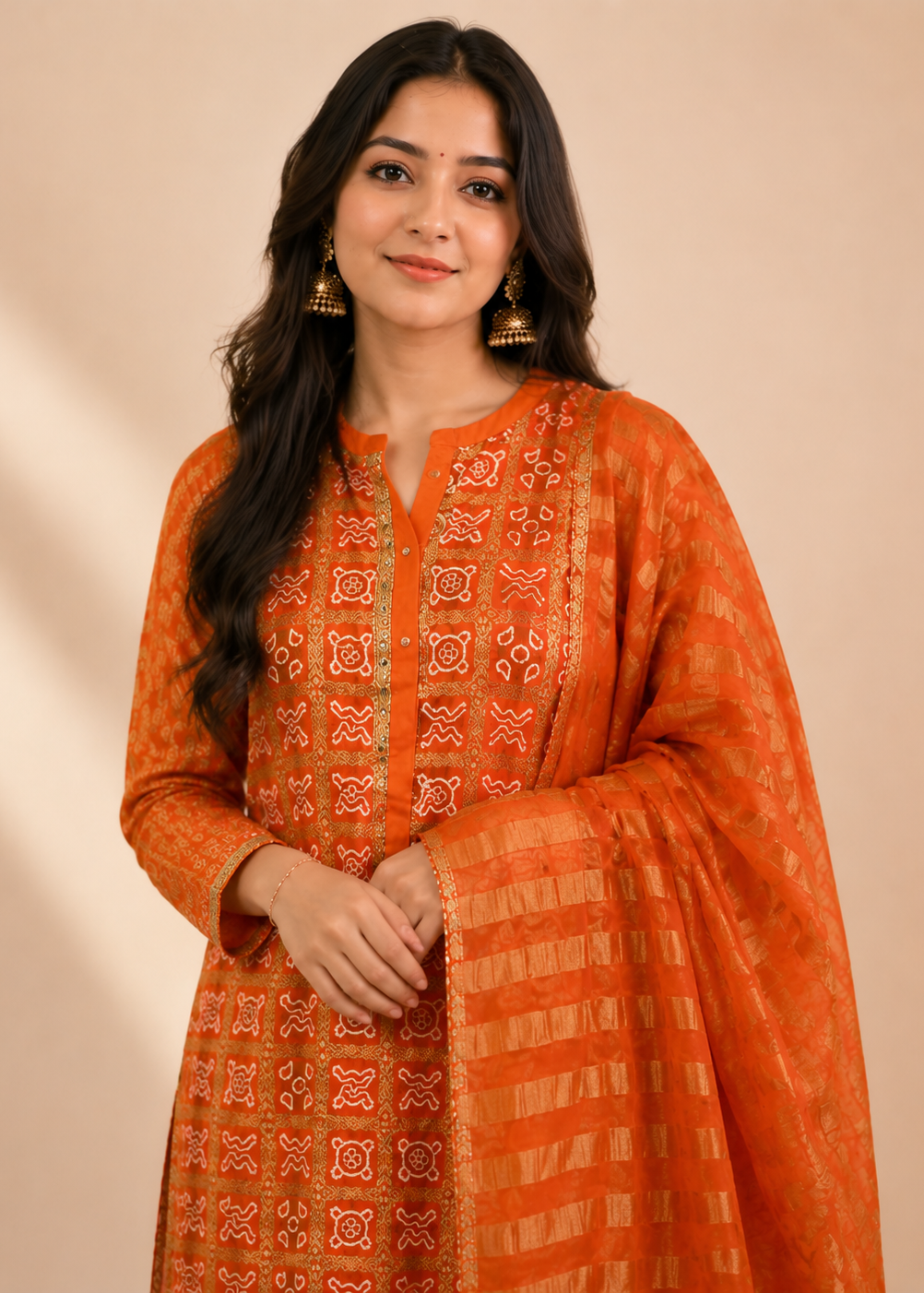 A woman modeling a bright orange traditional outfit with intricate red and gold embroidery, featuring a matching sheer dupatta draped over her arm, set against a solid beige background.