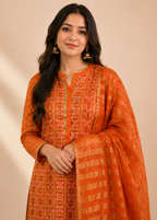 A woman modeling a bright orange traditional outfit with intricate red and gold embroidery, featuring a matching sheer dupatta draped over her arm, set against a solid beige background.