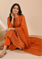 A woman models an orange traditional South Asian outfit consisting of a kurta with matching embroidered dupatta and wide-leg trousers, set against a warm beige background.