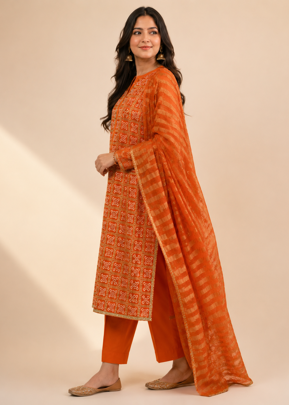 A woman modeling an orange traditional outfit with intricate embroidery on the kurta, matching orange pants, and a striped orange dupatta, set against a warm beige background.