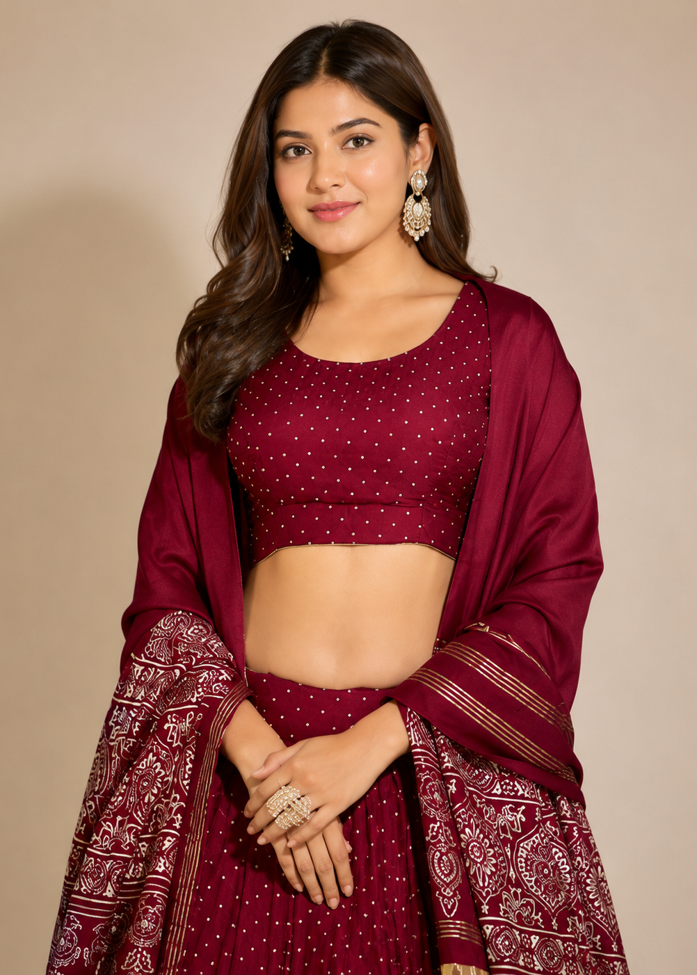 A woman modeling a deep maroon/burgundy traditional outfit with a matching dupatta, featuring a crop top, flared skirt, and intricate gold embroidery and border patterns, set against a plain beige studio background.