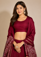 A woman modeling a deep maroon/burgundy traditional outfit with a matching dupatta, featuring a crop top, flared skirt, and intricate gold embroidery and border patterns, set against a plain beige studio background.