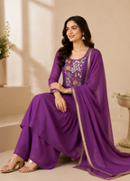 A woman models a purple ethnic salwar kameez suit set with floral embroidery on the chest and a matching sheer dupatta with gold border detailing, seated in a warm beige studio setting.