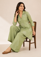 A woman models a sage green two-piece kurta and trouser set with white floral embroidery, seated on a wooden chair against a warm beige background.