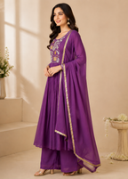 A woman models a purple traditional Indian outfit with a matching long dupatta, featuring gold zari borders and floral embroidery on the bodice, set against a warm beige background.