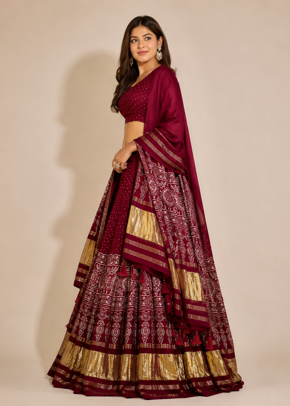 A woman modeling a traditional Indian lehenga choli set in deep maroon with intricate gold and silver metallic patterns, set against a plain beige studio background.