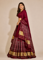A woman modeling a traditional Indian lehenga choli set in deep maroon with intricate gold and silver metallic patterns, set against a plain beige studio background.