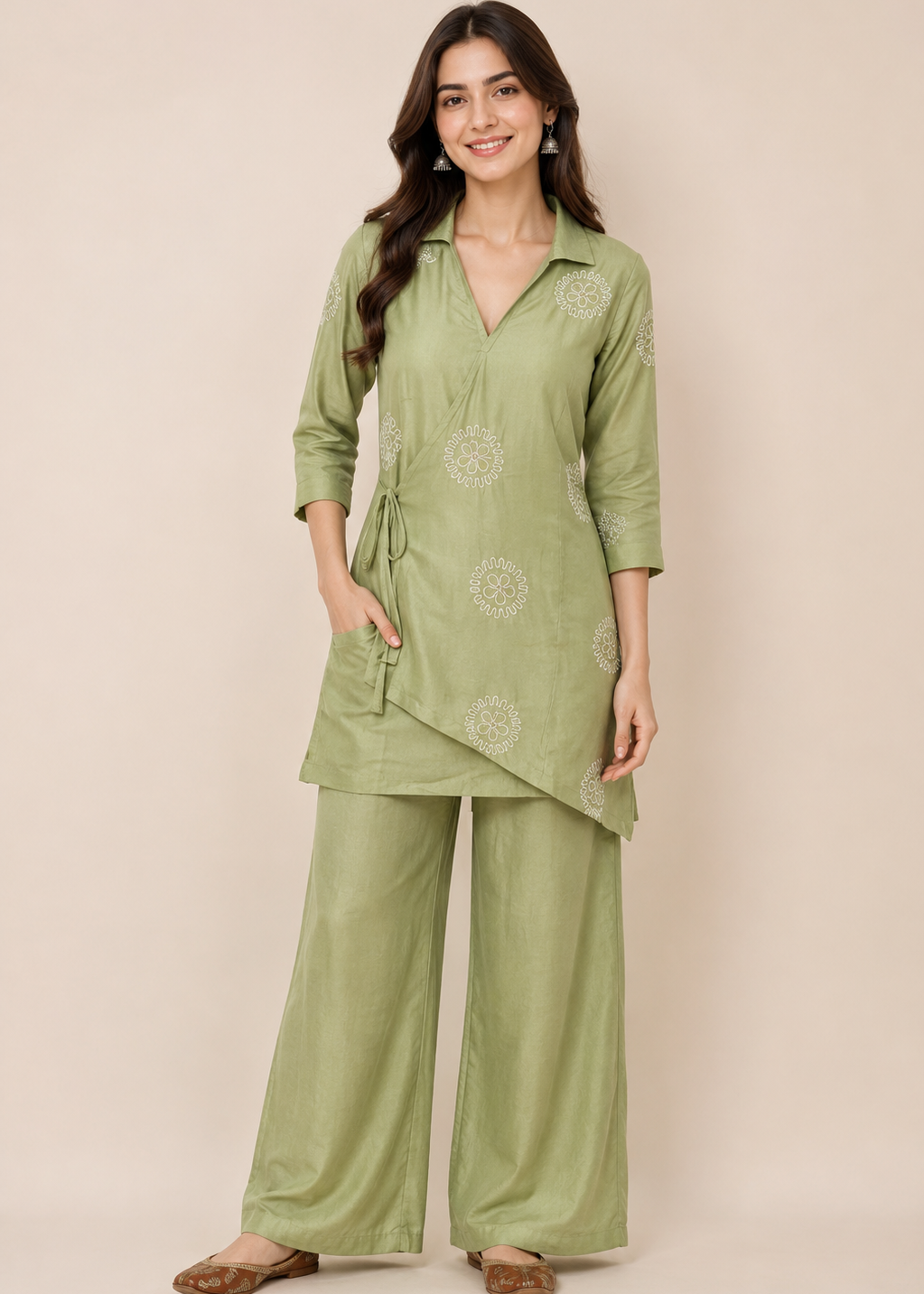 A woman modeling a sage green two-piece kurta set with matching wide-leg trousers, featuring light beige floral embroidery, against a solid beige studio background.