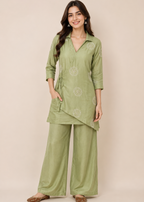 A woman modeling a sage green two-piece kurta set with matching wide-leg trousers, featuring light beige floral embroidery, against a solid beige studio background.