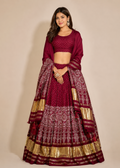 A woman modeling a traditional Indian lehenga choli set in deep maroon with intricate gold and silver zari embroidery and a matching dupatta, set against a plain beige studio background.