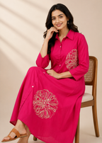 A woman wearing a bright pink long-sleeve maxi shirt dress with white floral embroidery, sitting on a wooden chair against a beige background with soft shadow patterns.