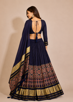 A woman modeling a navy blue traditional Indian lehenga choli set with intricate gold, pink, and red patterns on the skirt, shown from the back against a solid beige studio background.