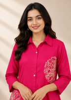 A woman wearing a bright pink button-up tunic dress with white floral embroidery on the chest and lower skirt, set against a warm beige background.