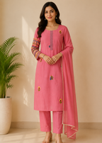 A woman modeling a pink traditional Indian salwar kameez set with matching pants and dupatta, featuring colorful embroidered details on the sleeves and front, set against a warm beige wall with a potted plant on the left.