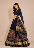 A woman modeling a traditional Indian lehenga choli set in navy blue and gold. The skirt features intricate geometric and floral patterns in pink, gold, and navy, with a matching navy and gold dupatta draped over her shoulder.