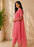 A woman models a pink Indian ethnic outfit consisting of a kurta, trousers, and a dupatta. The kurta features gold buttons and colorful striped embroidery on the sleeves, paired with matching pink trousers and a sheer pink dupatta, set against a warm beige wall with soft window shadows.