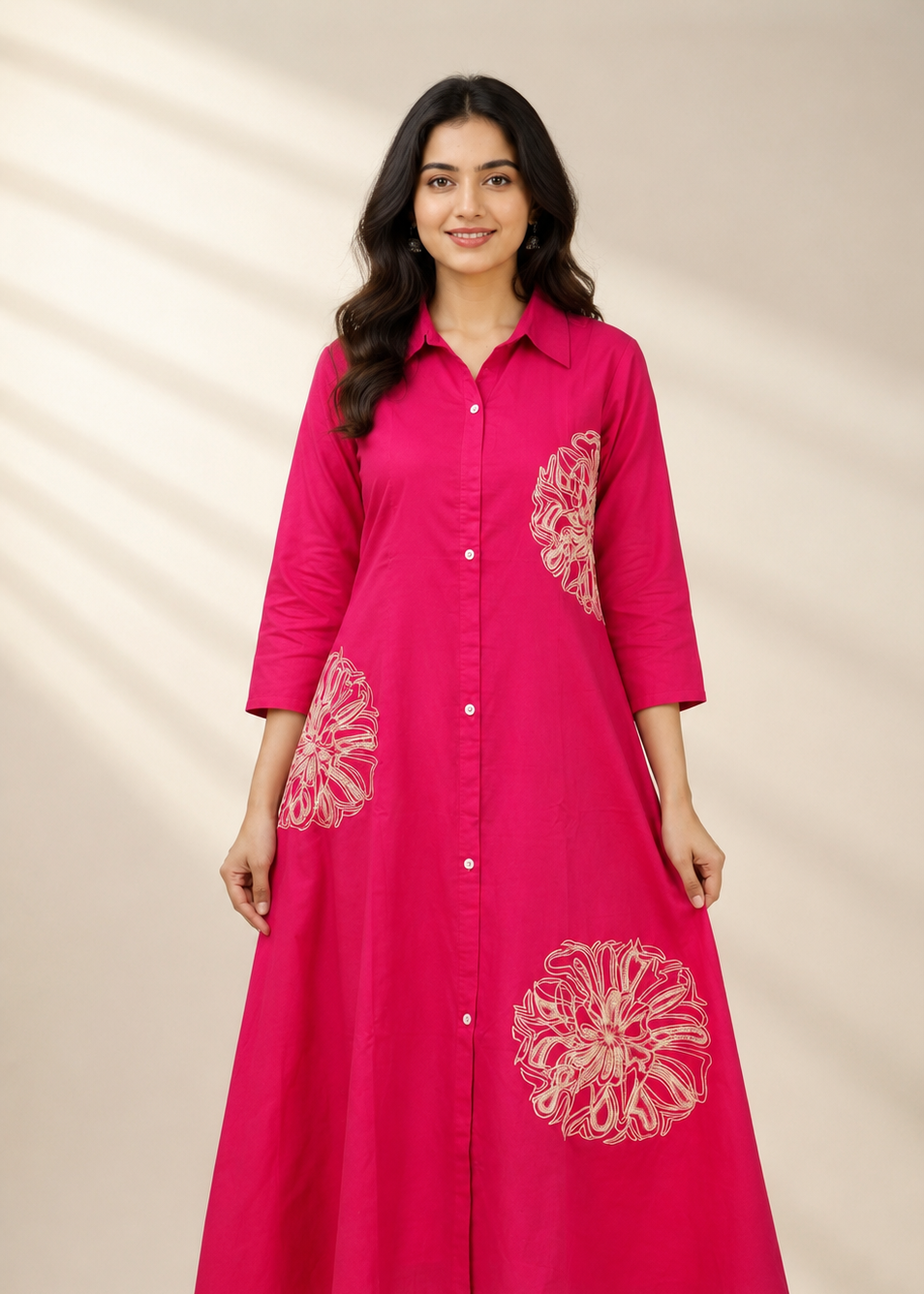 A woman modeling a bright pink long-sleeve button-up dress with white floral embroidery on the front and sleeves, set against a warm beige background with soft leaf shadows.