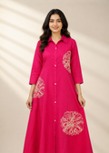 A woman modeling a bright pink long-sleeve button-up dress with white floral embroidery on the front and sleeves, set against a warm beige background with soft leaf shadows.