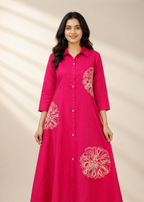 A woman modeling a bright pink long-sleeve button-up dress with white floral embroidery on the front and sleeves, set against a warm beige background with soft leaf shadows.