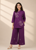 A woman modeling a purple two-piece kurta set with white floral embroidery, standing against a beige background with soft window shadow patterns.