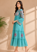 A woman modeling a tiered turquoise blue maxi dress with colorful embroidered details on the neckline, sleeves, and lower skirt, set against a warm beige wall with soft window shadows.