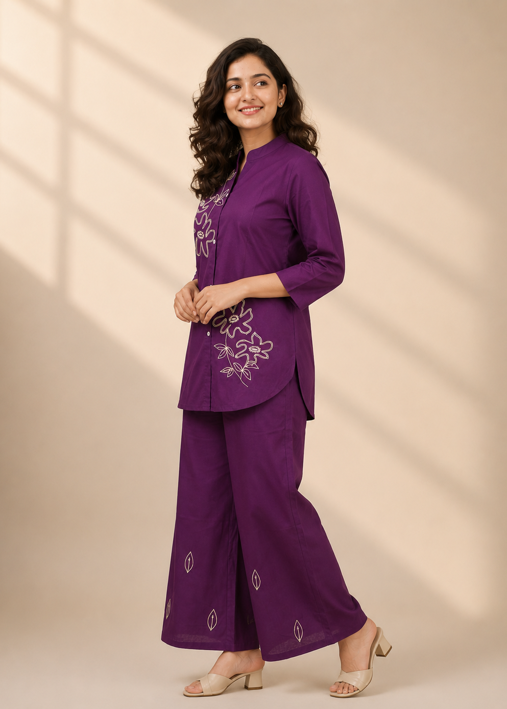 A woman modeling a purple two-piece kurta set with white floral embroidery, standing against a beige background with soft window shadow patterns.
