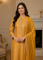 A woman modeling a traditional Indian outfit consisting of a mustard yellow embroidered kurta and a matching dupatta, set against an elegant indoor background with a fireplace.